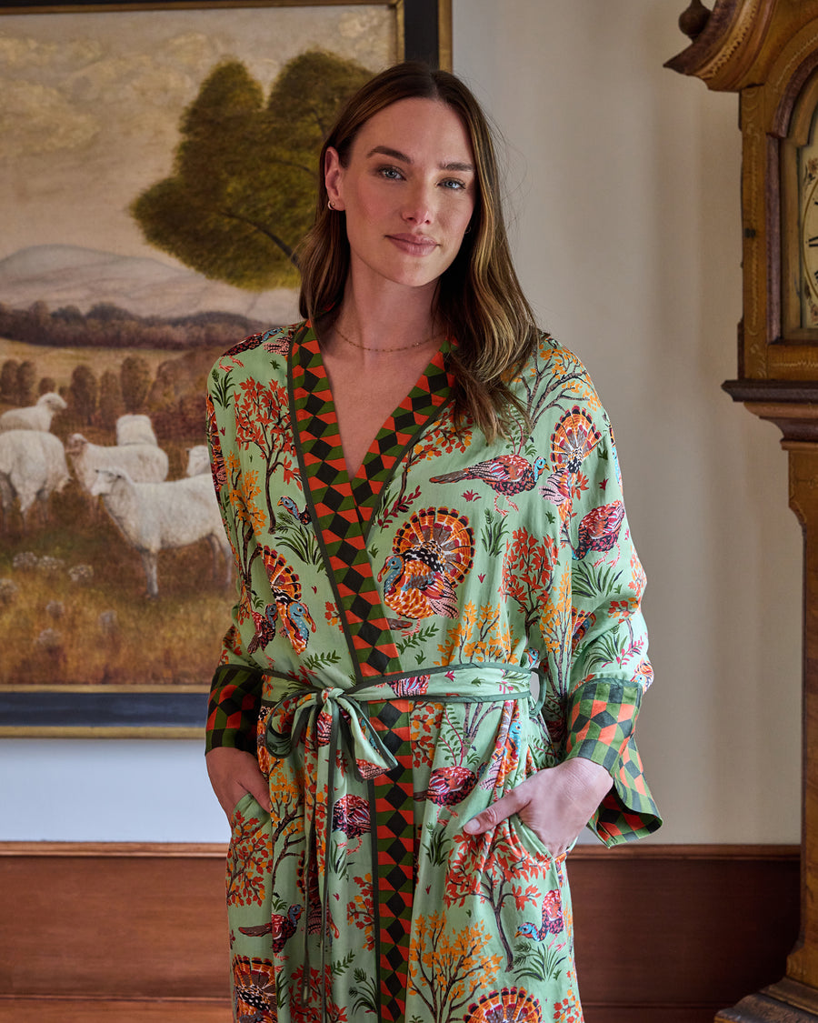 Person wearing a soft sage Turkey Trot long robe with turkey and autumn foliage print, contrasting geometric trim, belt, and pockets.