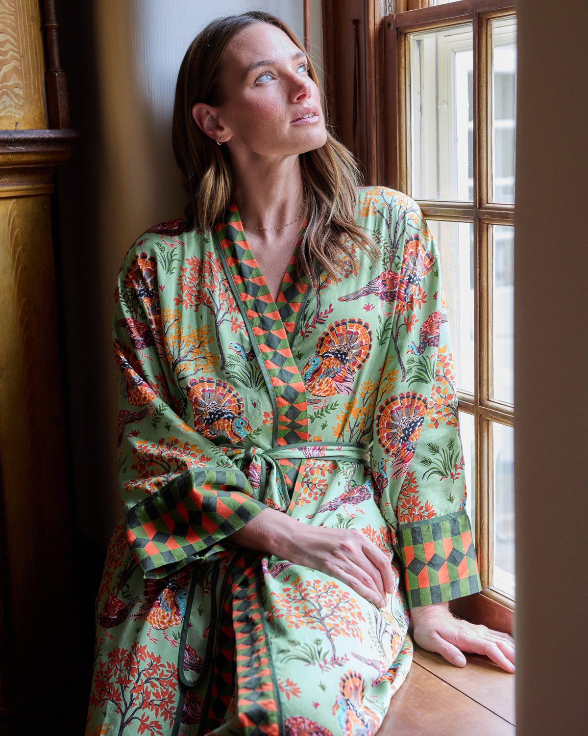 Person seated by a window wearing a soft sage Turkey Trot long robe with turkey and autumn foliage print and contrasting geometric trim.