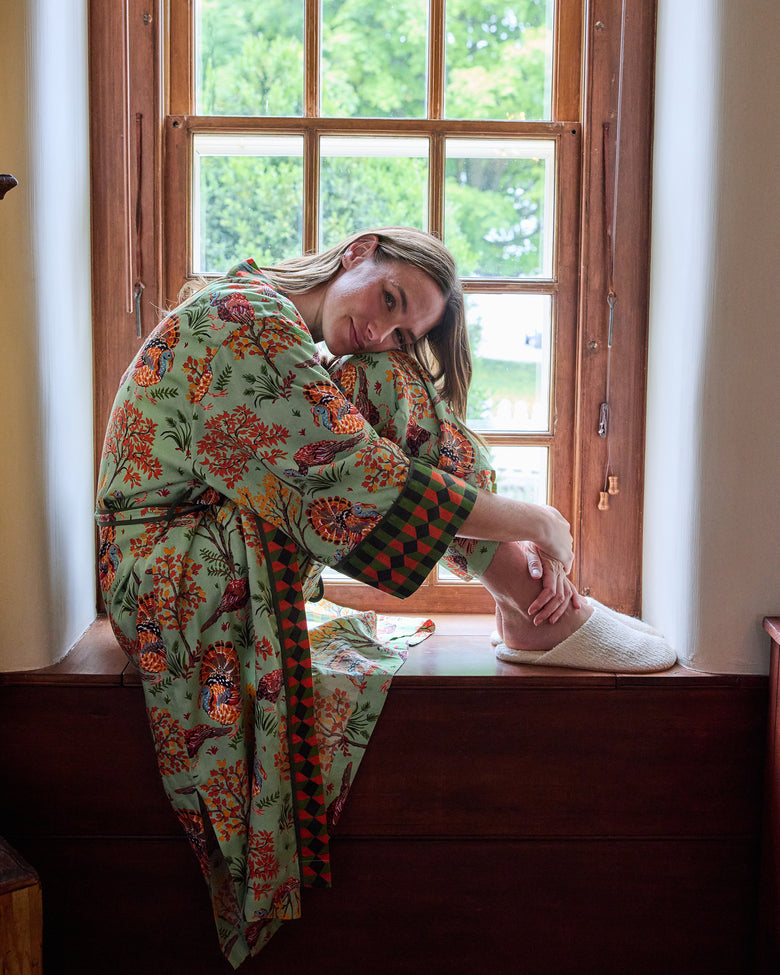 Person wearing a sage robe with a colorful turkey and autumn foliage print, seated by a wooden window.