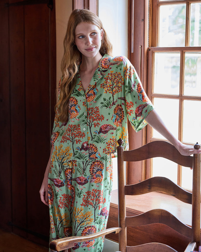 Person wearing a sage button-front pajama set with a colorful turkey and autumn foliage print, standing beside a wooden chair.
