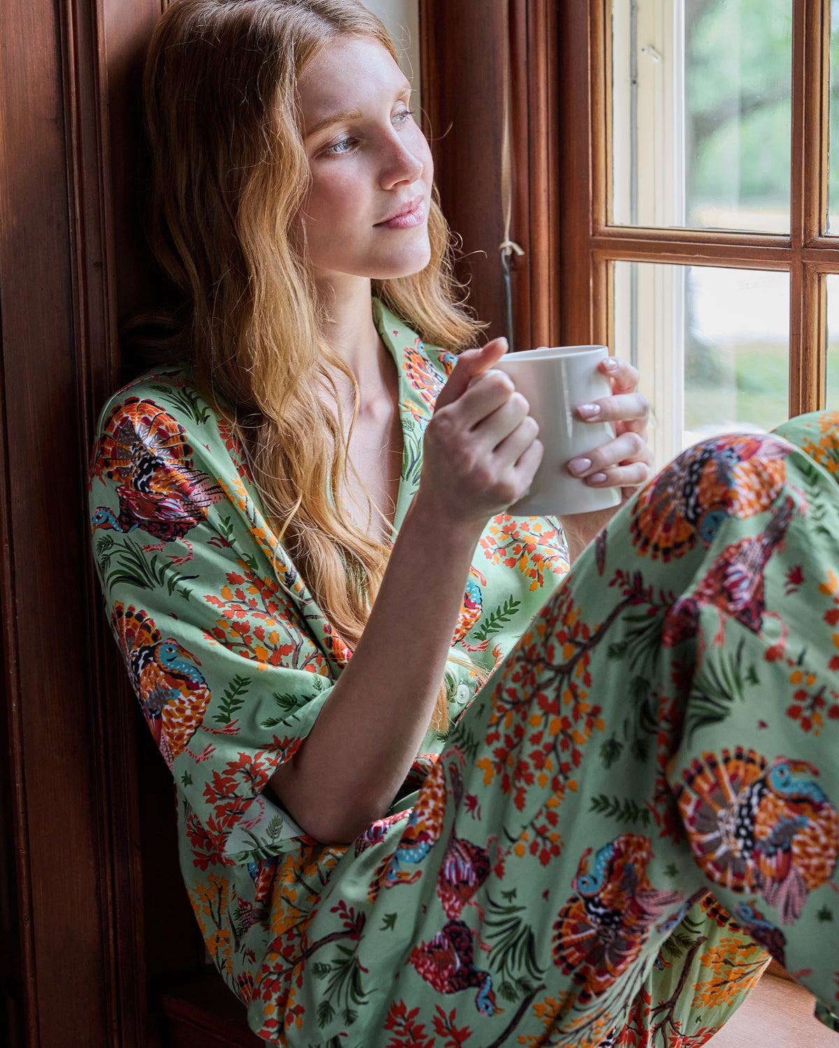 Person wearing a sage turkey-print pajama set and holding a white mug while seated beside a window.