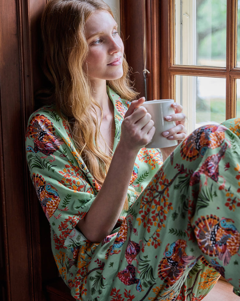 Person wearing a sage turkey-print pajama set and holding a white mug while seated beside a window.