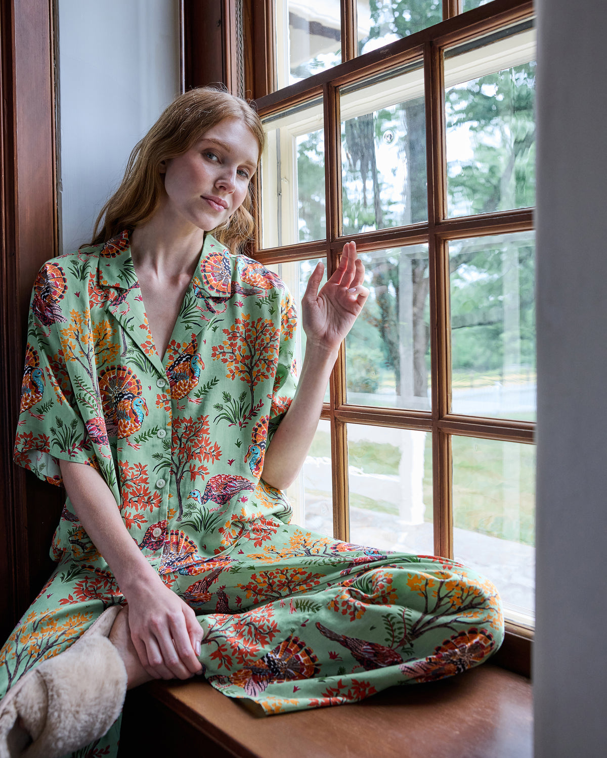 Person wearing a sage pajama set with a colorful turkey and autumn foliage print, seated by a window.