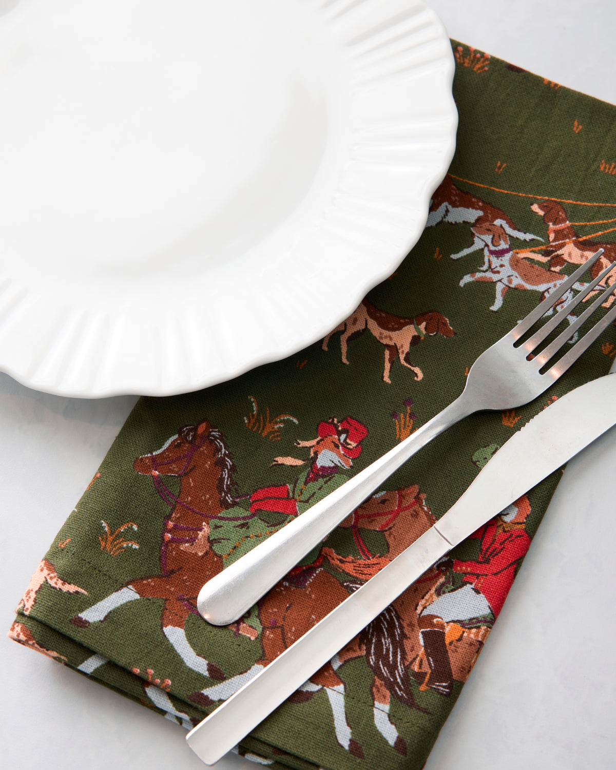 Close-up of a Fox & Hounds dinner napkin beneath a white plate, fork, and knife, showing the horse-and-hounds print.