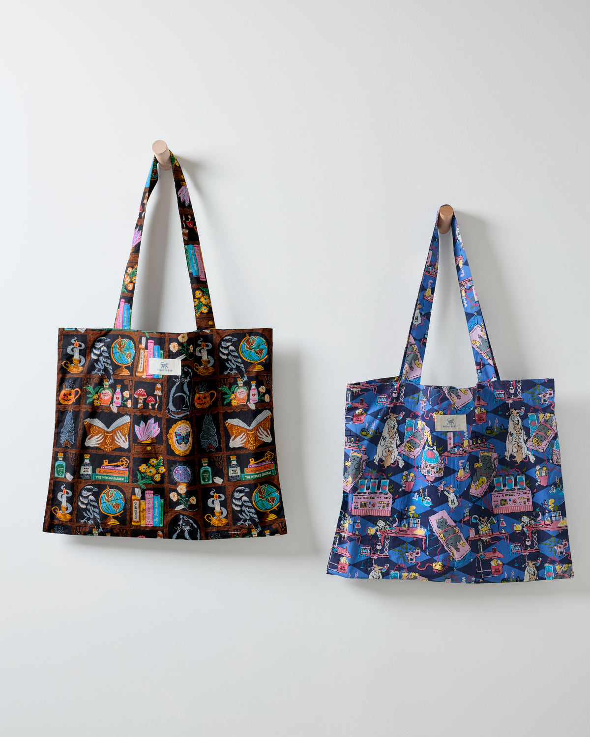 Two Halloween Best printed tote bags hanging on a wall, one dark It's Alive print and one blue-purple Enchanted Study print.