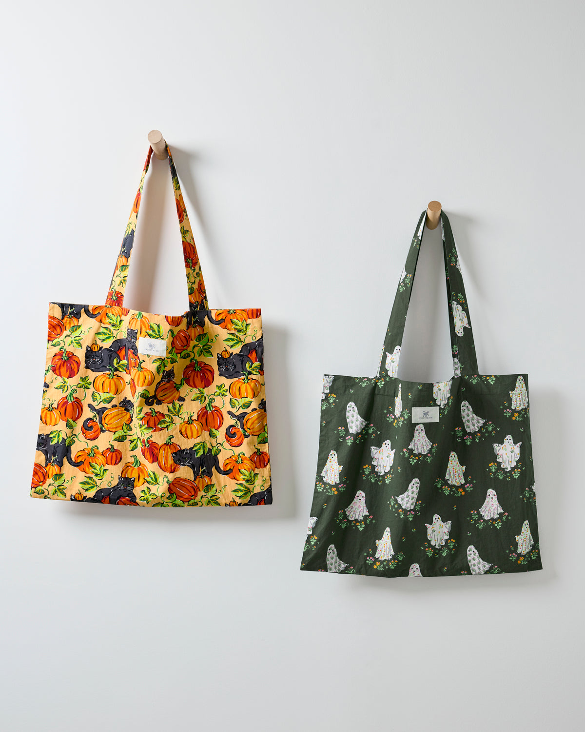 Two Halloween Garden printed tote bags hanging on a wall, one pumpkin-and-black-cat print and one Gardening Ghosts print.