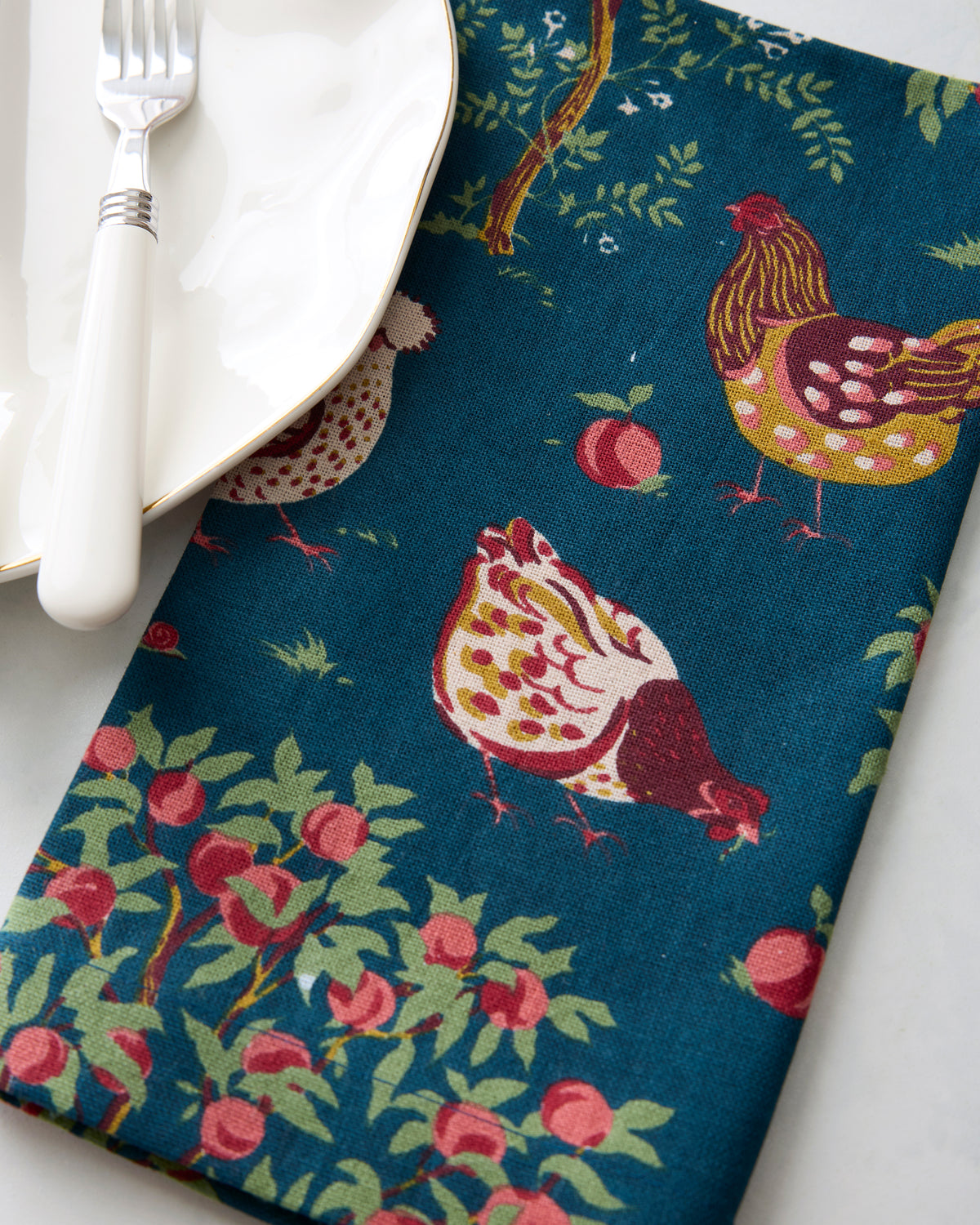 Close-up of the navy Hen House napkin print showing chickens, apples, and leafy trees beside a plate and fork.