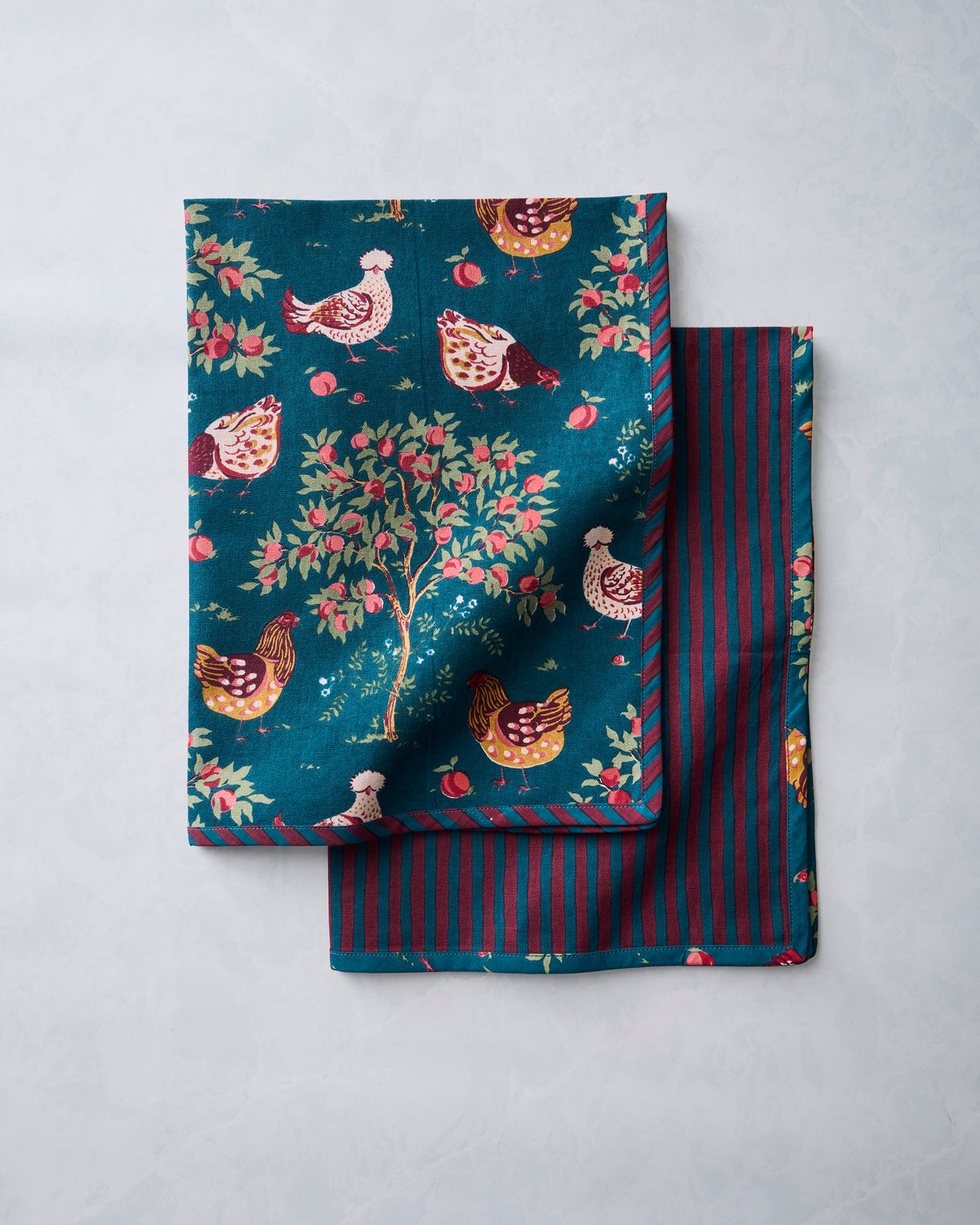 Two overlapping tea towels showing the chicken-and-apple-tree print and the dark teal and burgundy striped reverse.
