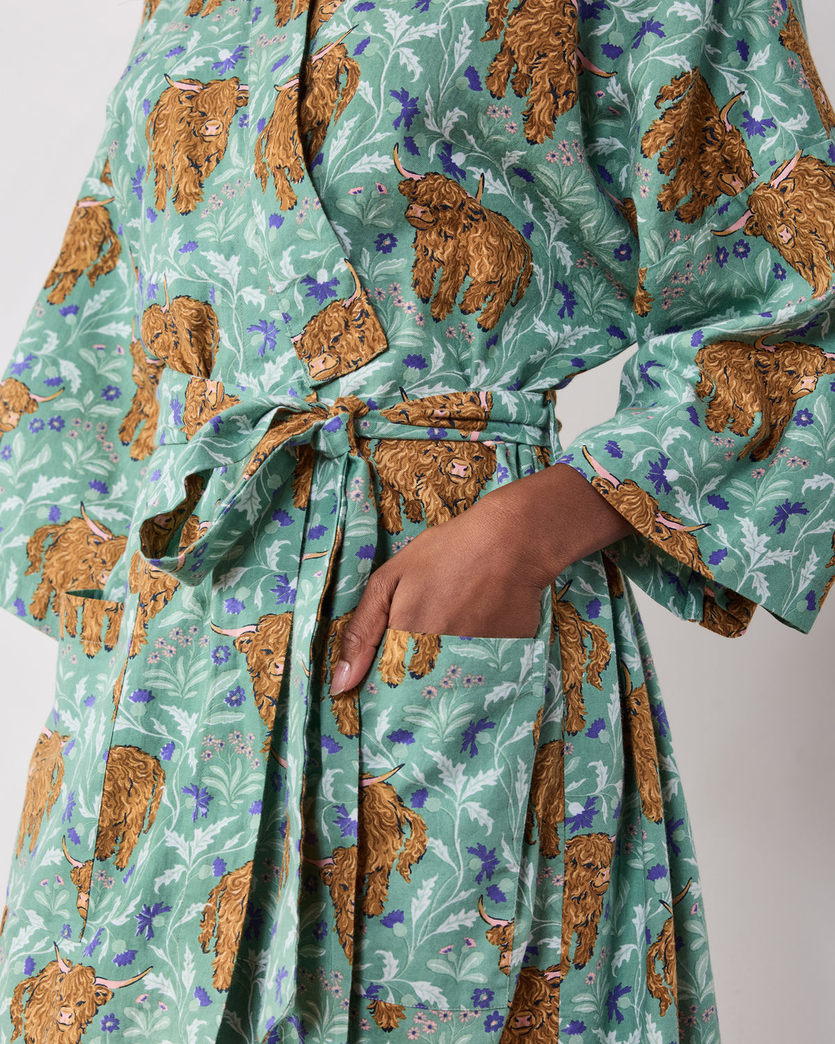 Close-up of the green Highland Cows robe showing the tied belt, wide sleeve, and hand in a front pocket.