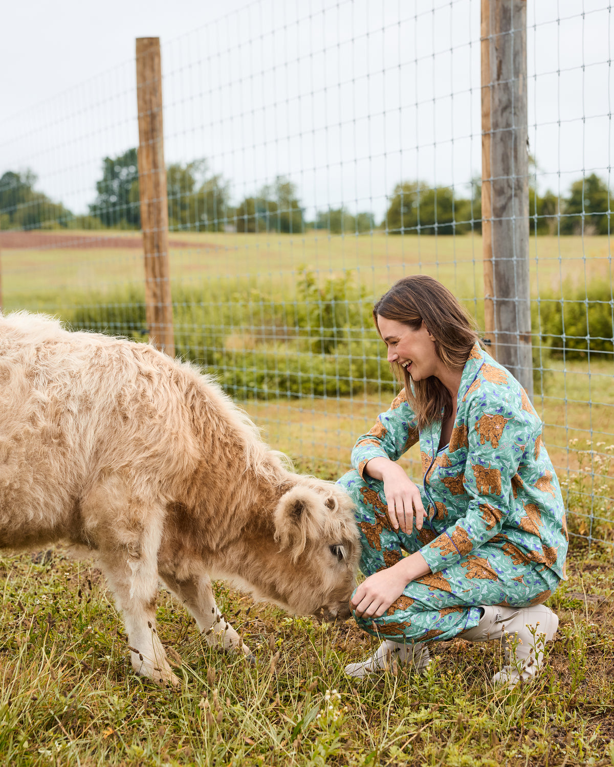Person wearing the Highland Cows printed long pajama set while crouching beside a Highland cow in a fenced field
