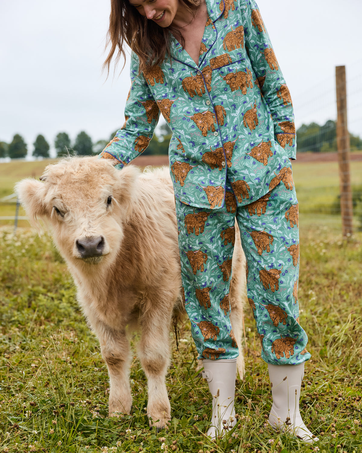 Person wearing a green Highland Cow print long-sleeve pajama set beside a calf in a field.