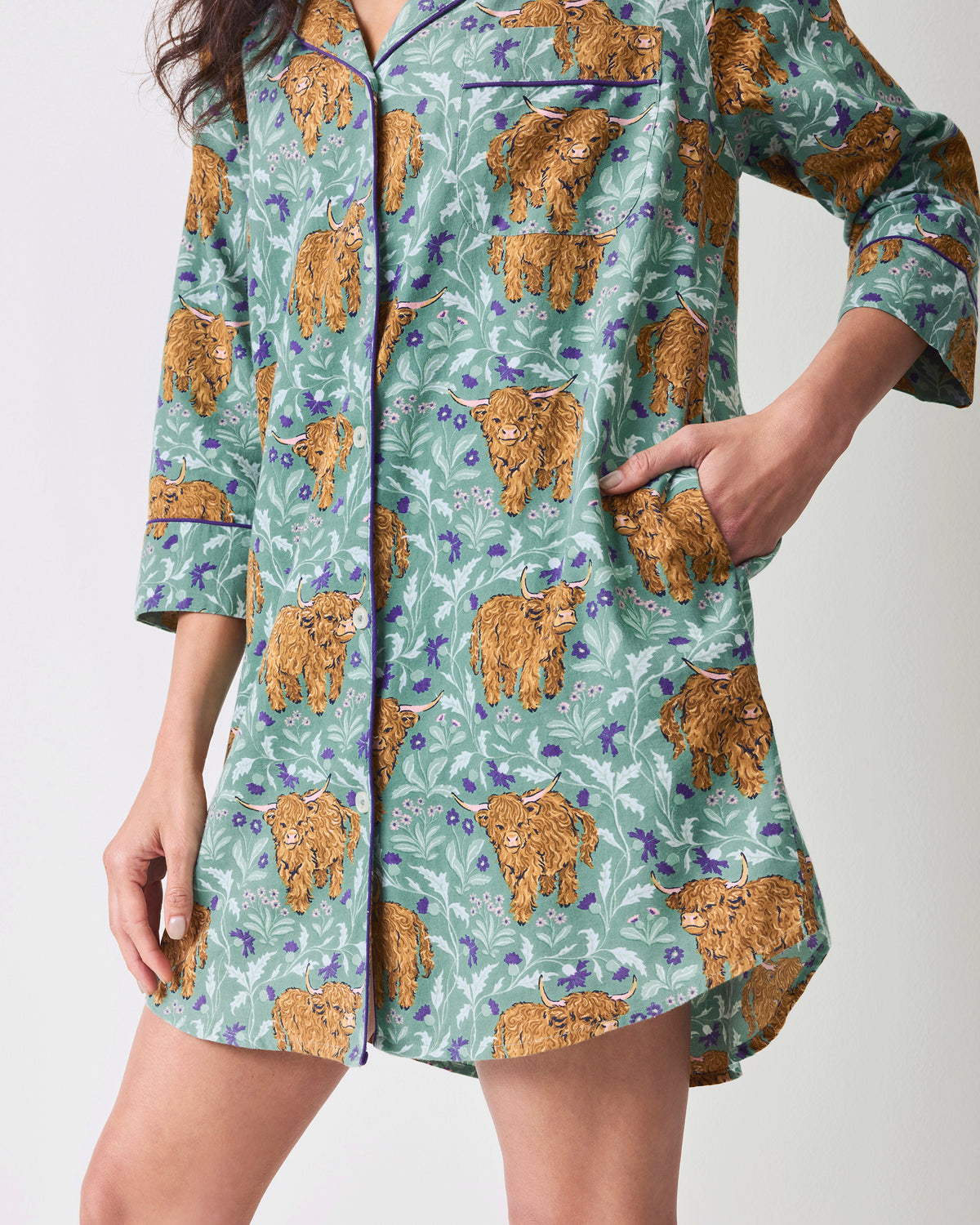 Close view of the lower front of a green Highland cow-print sleep shirt, including its pocket and curved hem.