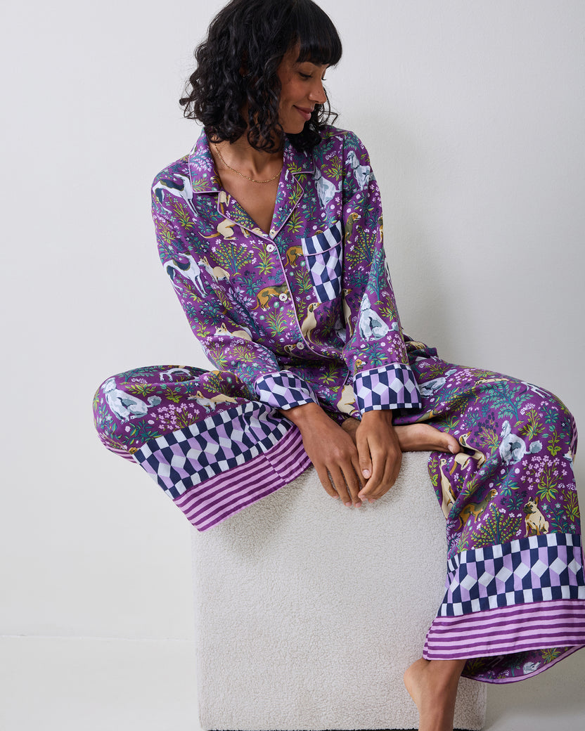 Person seated wearing a purple dog-and-floral print pajama set with a collared button-front top and wide-leg pants with geometric cuffs and striped hems.