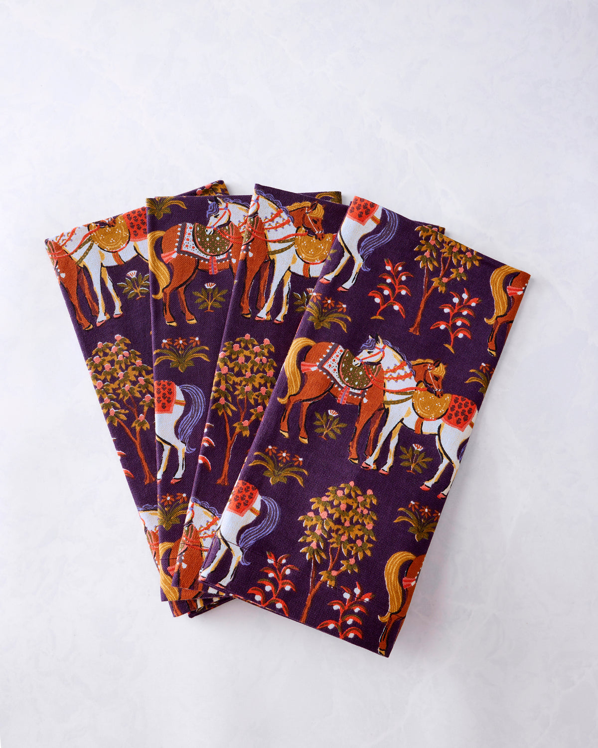 Four purple dinner napkins fanned out, printed with horses, trees, and floral motifs.
