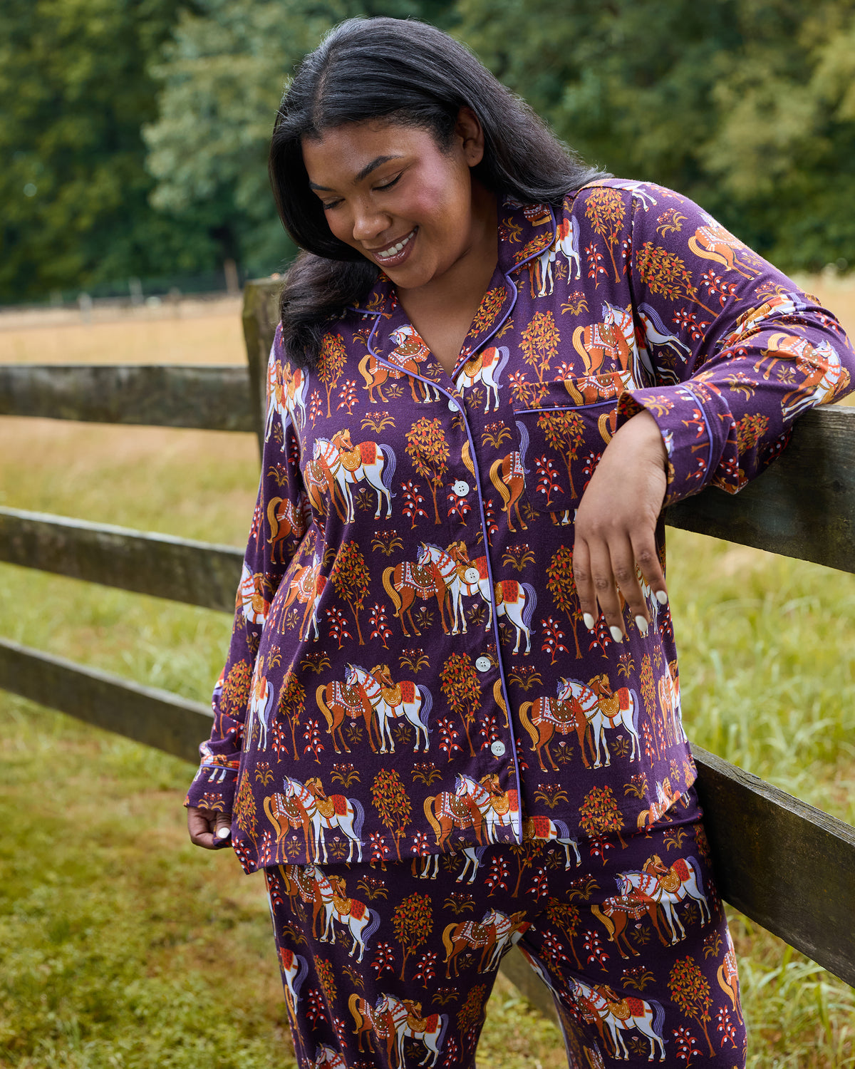 Person wearing the long-sleeve Queen’s Horses pajama set with horse-and-tree print, leaning against a wooden fence outdoors.