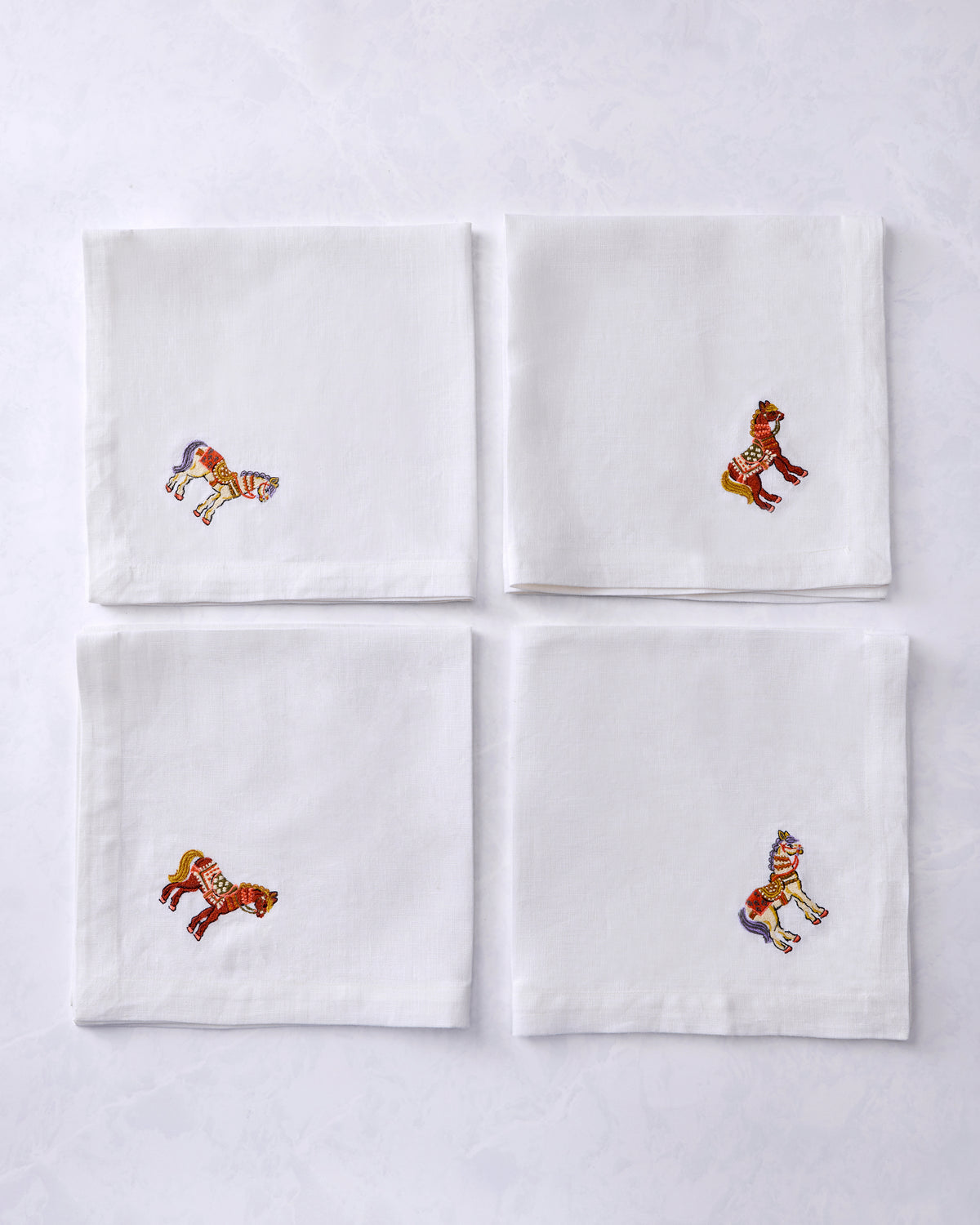 Four white dinner napkins arranged in a square, each with an embroidered horse in a different pose.