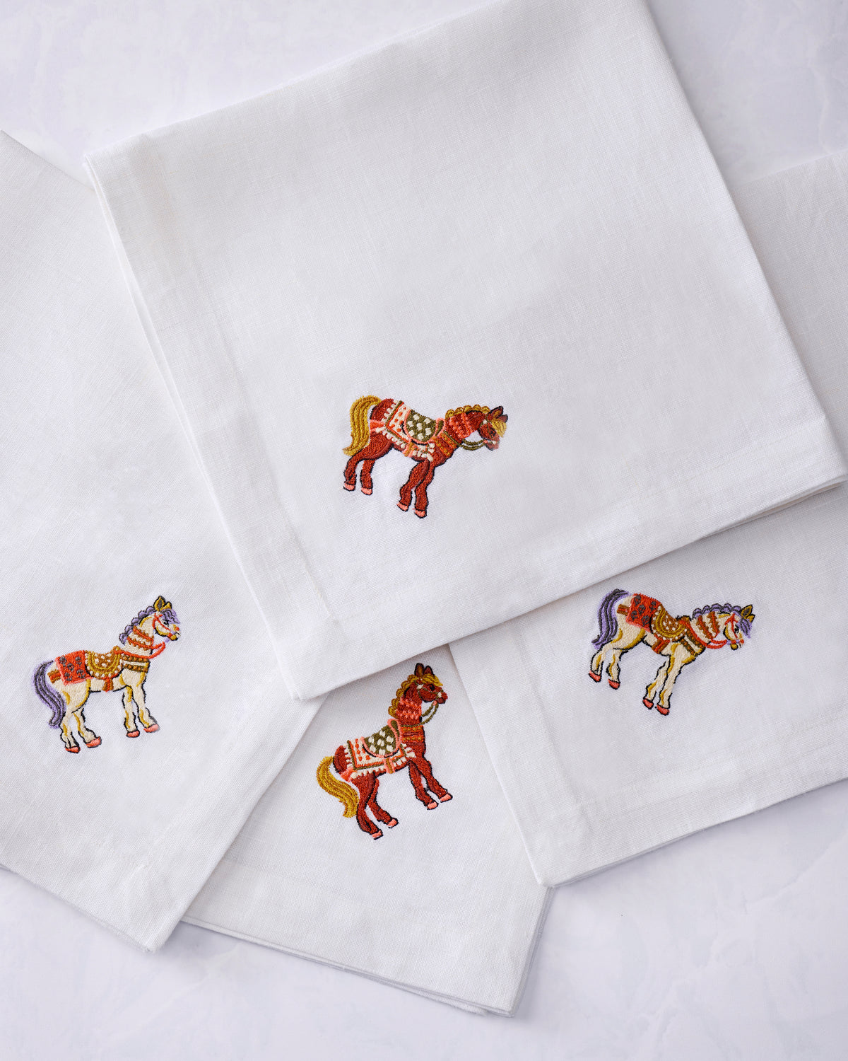 Close-up arrangement of four white dinner napkins, each embroidered with a colorful horse.