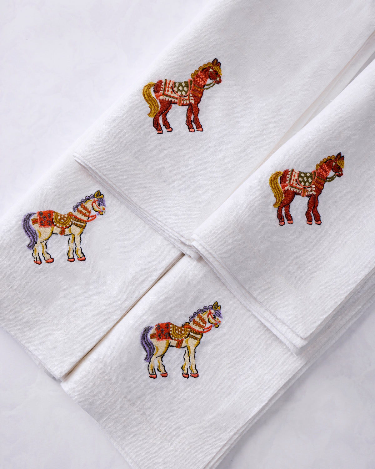 Close-up arrangement of four white dinner napkins with colorful embroidered horse designs.