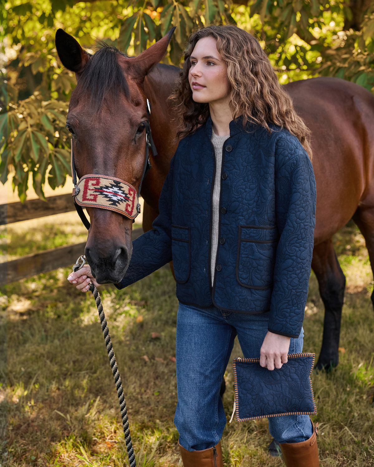 Close view of a person wearing a navy quilted jacket beside a brown horse and holding a matching pouch.