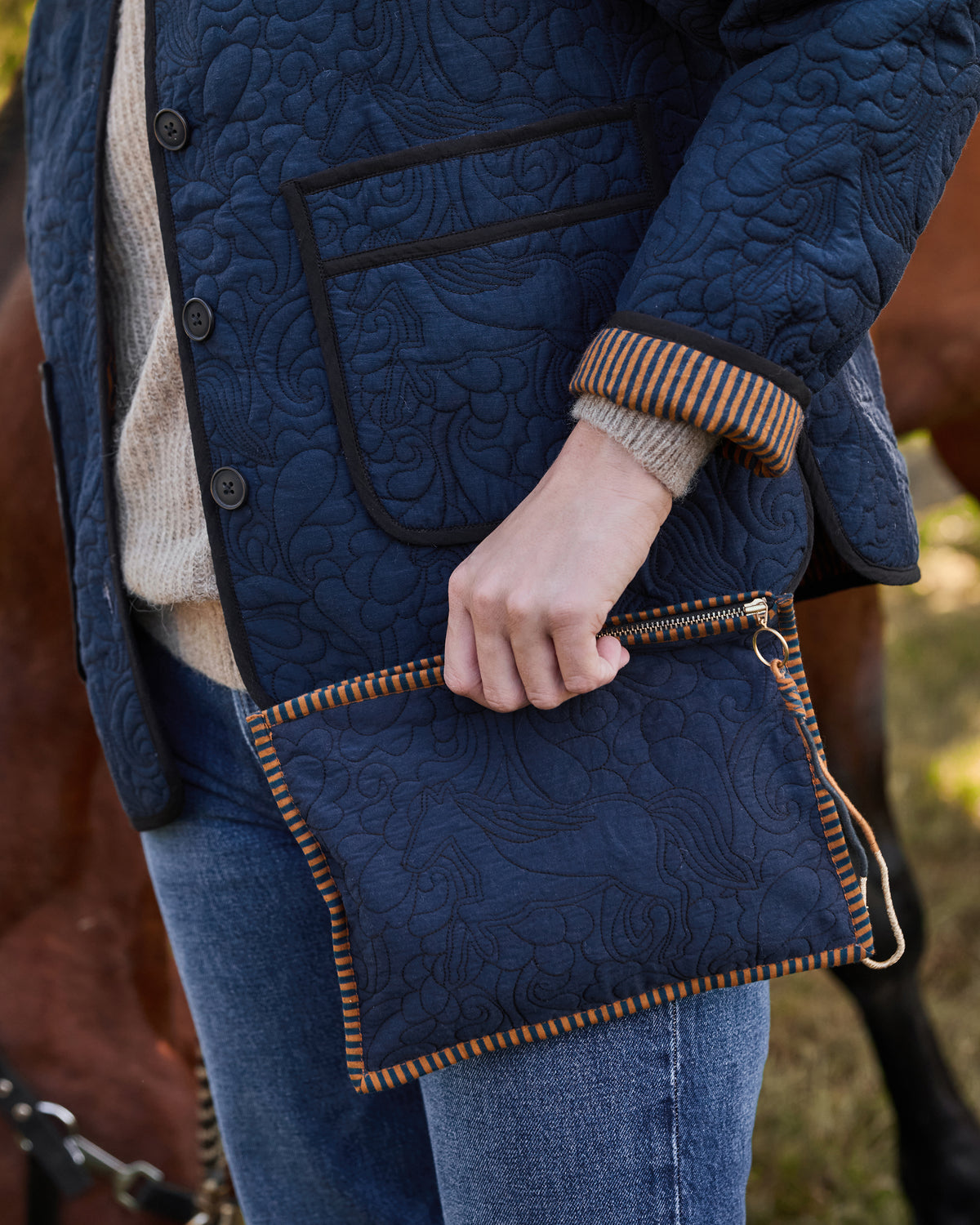 Close-up of the Vintage Indigo Marley Pouch held against a quilted equestrian jacket, showing its horse motif, striped trim, zipper, and wrist strap.