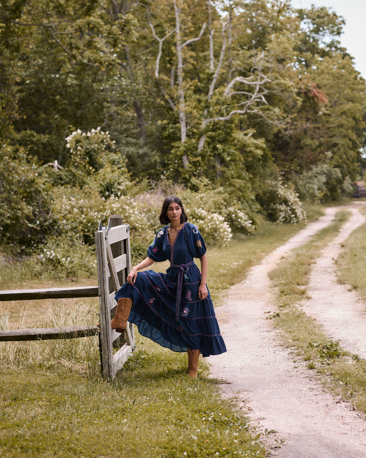 Model wearing the navy To and From maxi dress with a tied waist belt and embroidered motifs beside a garden fence.
