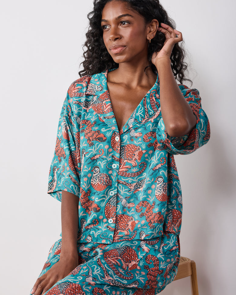 Seated model wearing the teal Southwestern Critters satin Wildest Dreams set, showing a short-sleeve button-front top and matching pants.