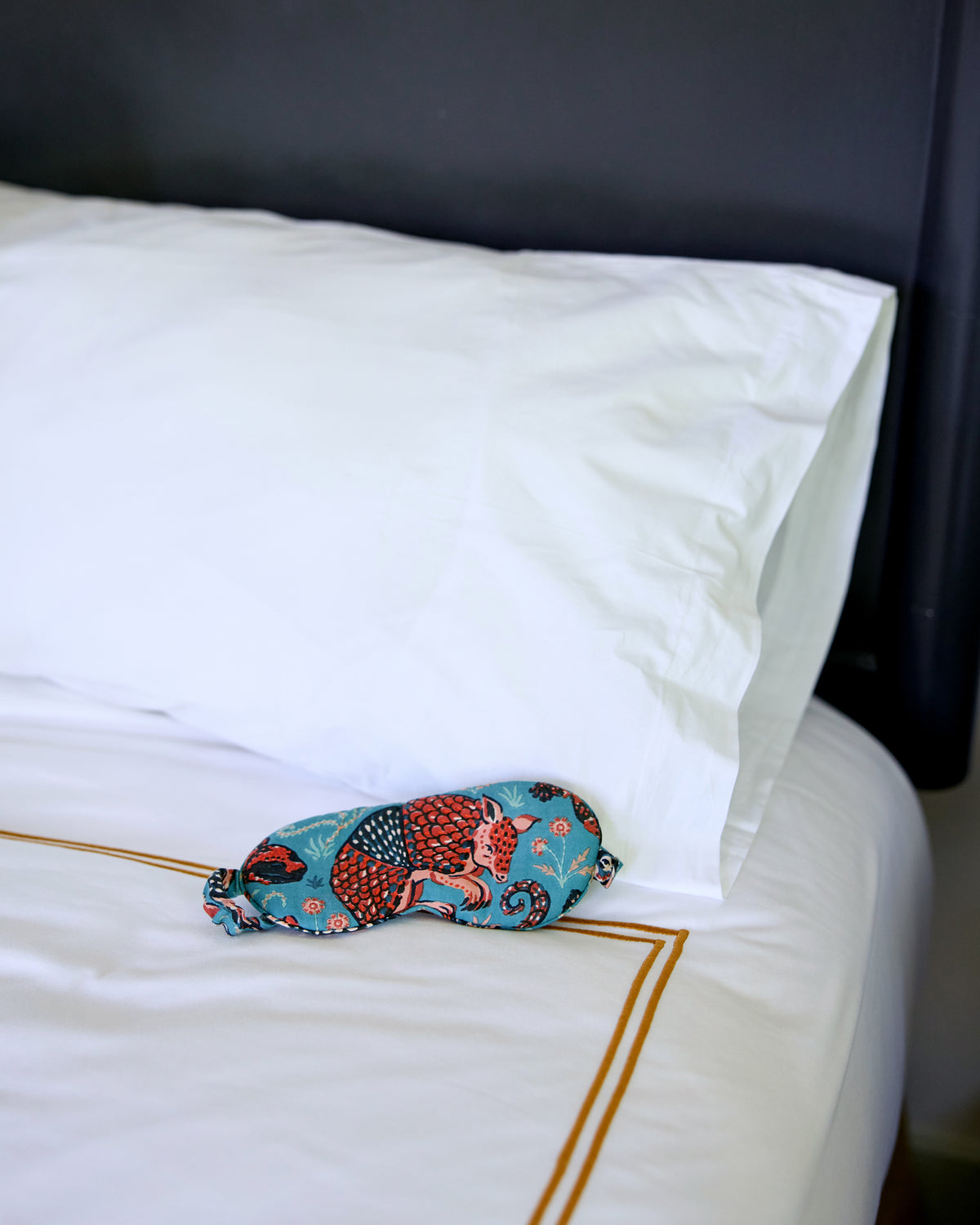 Teal Canyon Critters contoured eye mask resting on a white bed, with an adjustable gathered strap and a red armadillo print.