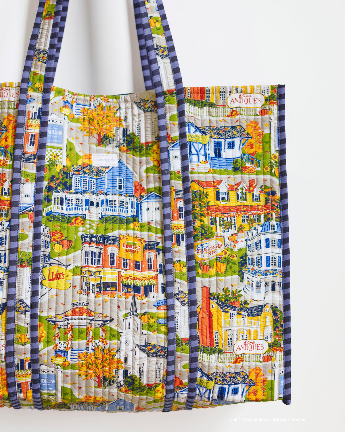 Close-up of the Stars Hollow Traveler Tote’s quilted illustrated town print, showing storefronts, houses, pumpkins, and blue plaid trim.