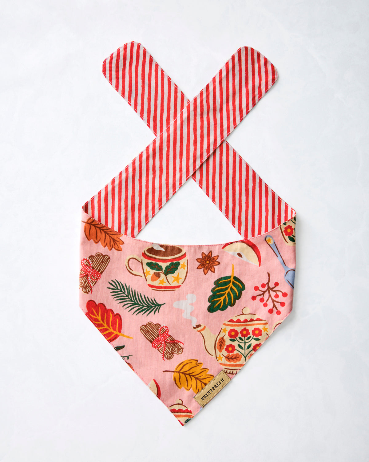 Pet bandana displayed flat with a pink tea-and-autumn print, red-and-white striped tie straps, and a pointed lower edge.