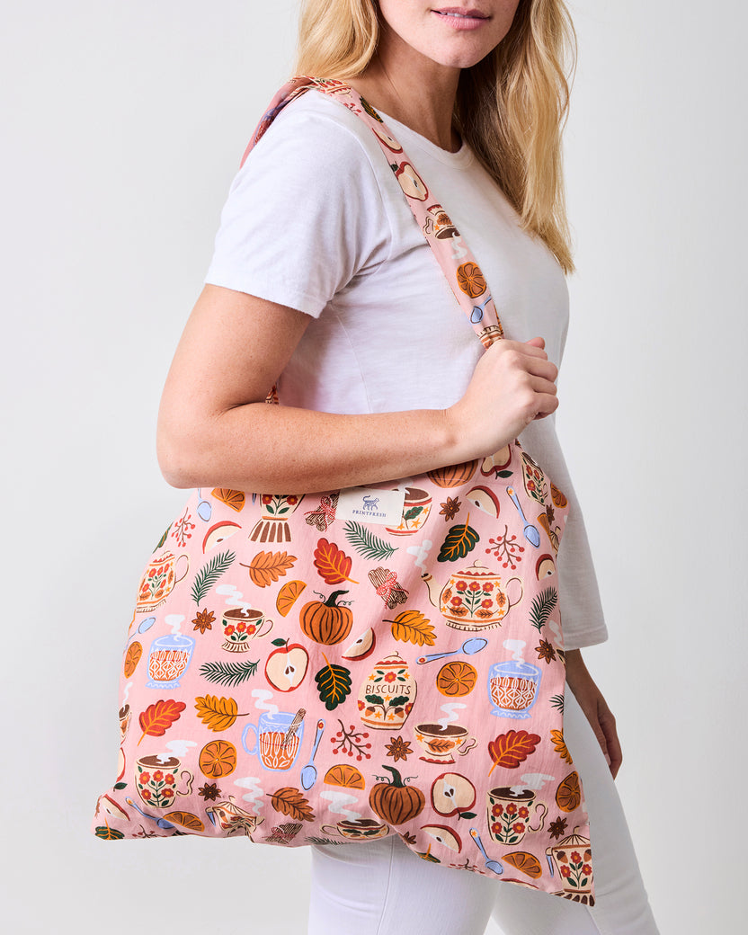 Pink tote bag with shoulder straps and a printed pattern of tea cups, leaves, fruit, and autumn motifs, shown over a white shirt.