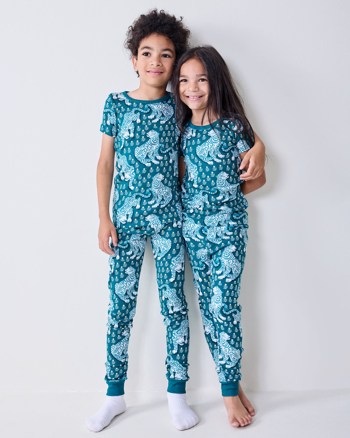 Two children stand together wearing matching teal tiger-print short-sleeve pajama sets.