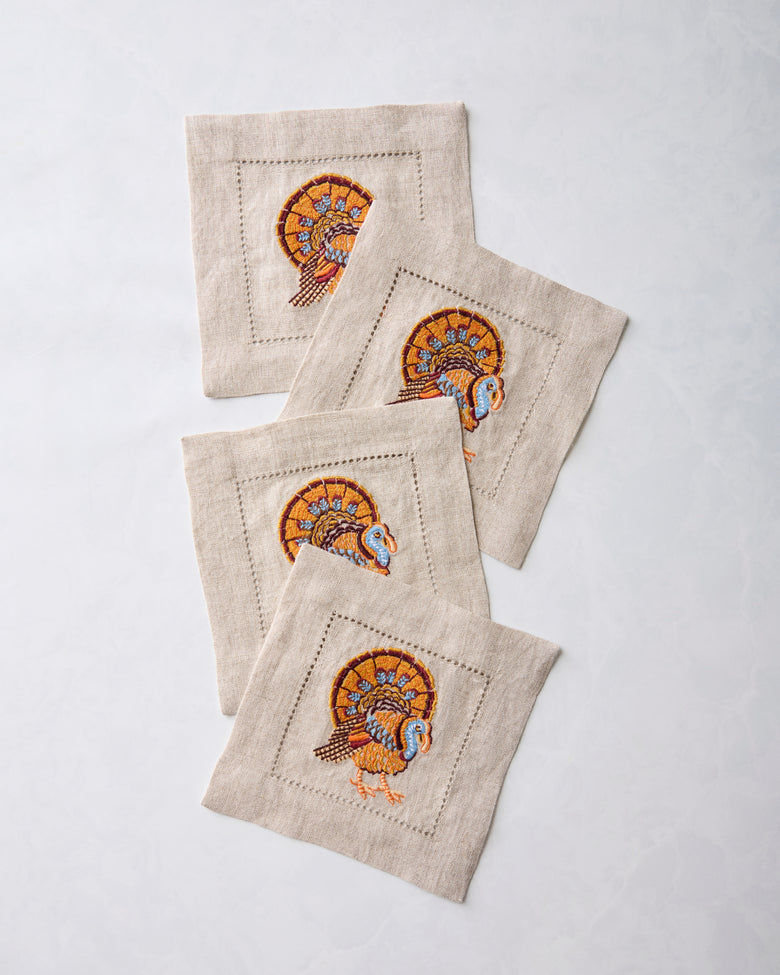 Four natural cocktail napkins with embroidered turkey motifs arranged in an overlapping stack.