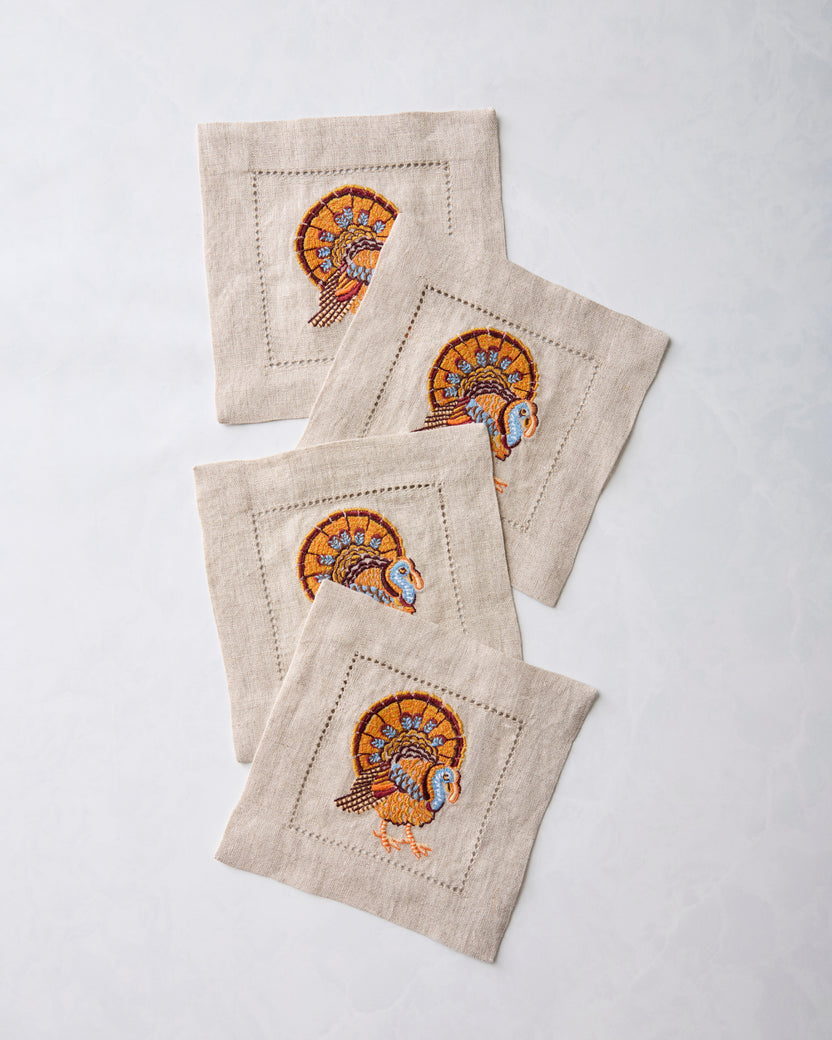 Four natural cocktail napkins with embroidered turkey motifs arranged in an overlapping stack.