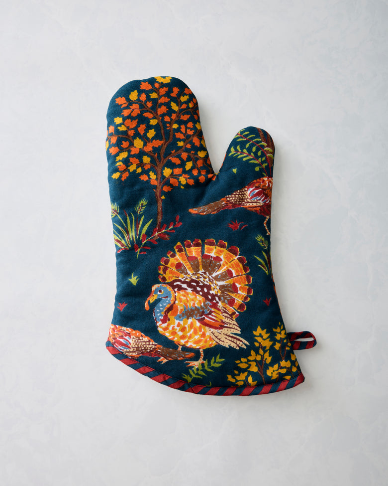 Navy oven mitt with a colorful turkey, tree, leaf, and grass print; hanging loop visible at the side.