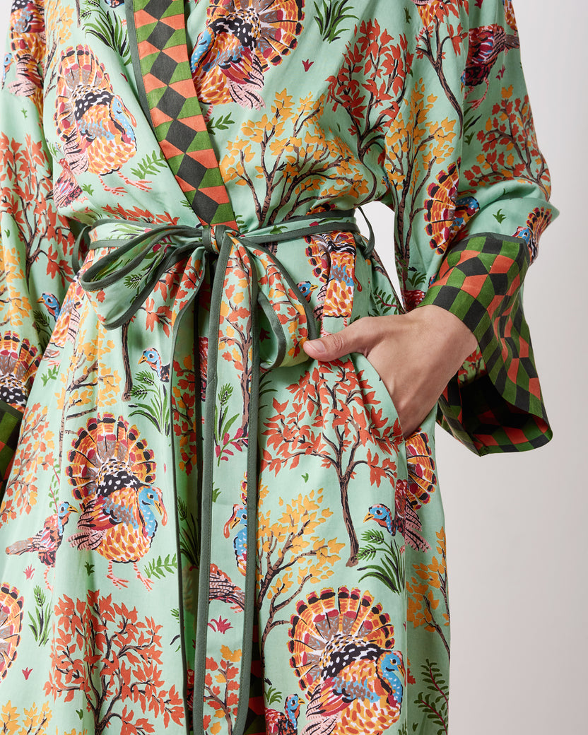Close-up of a soft sage Turkey Trot robe showing the turkey-and-autumn-tree print, tied waist belt, pocket, and contrasting diamond-patterned cuff.