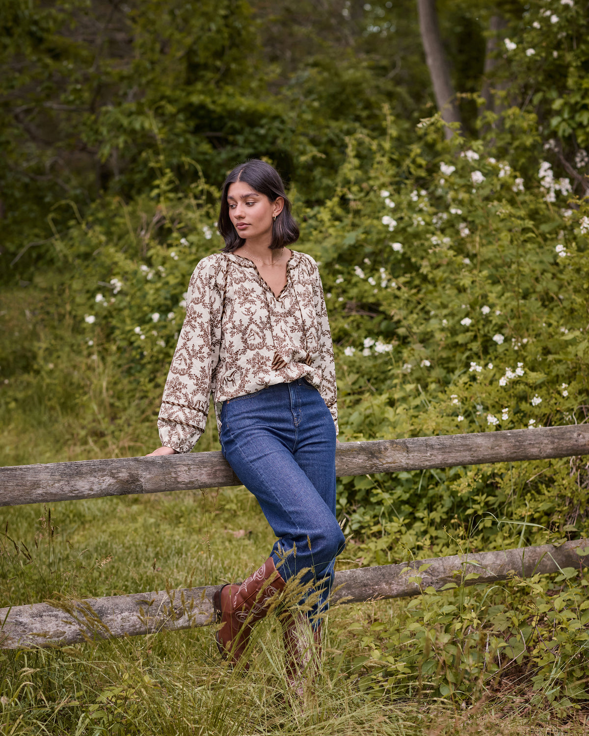 Person wearing the oat Western Crown Kaia blouse with horse, floral, and horseshoe print while sitting on a wooden fence outdoors.