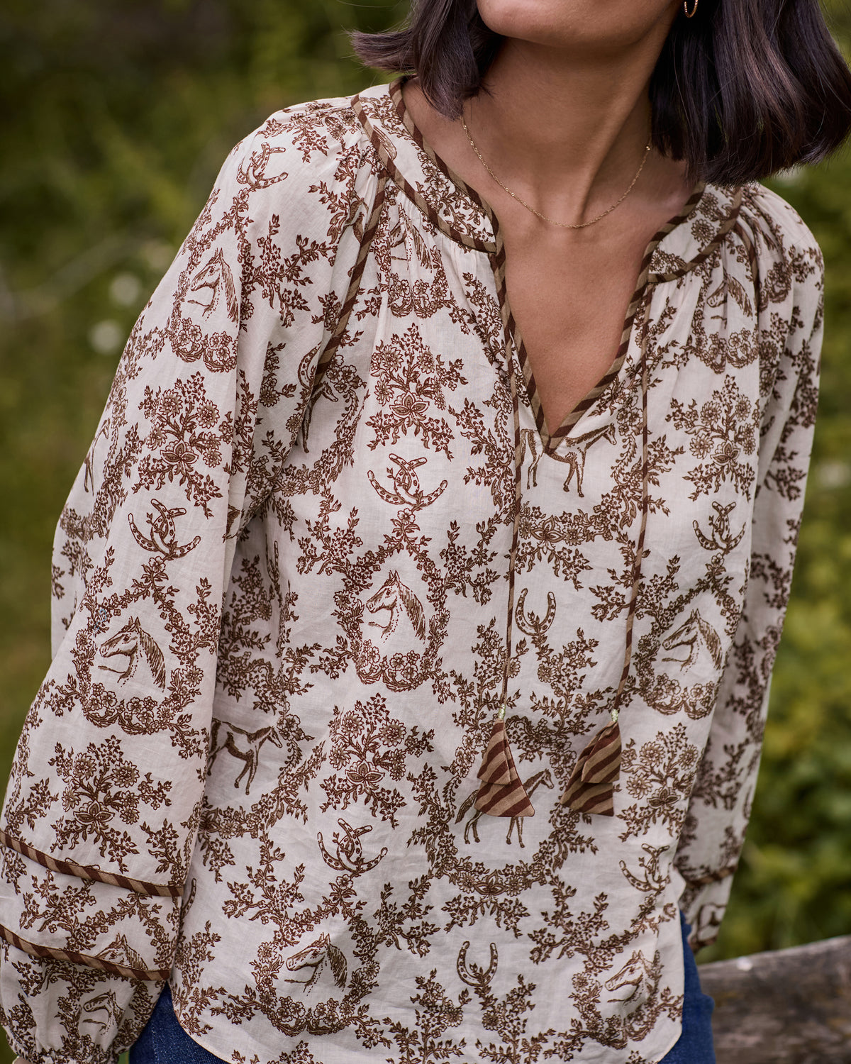 Close-up of the oat Western Crown Kaia blouse showing its horse, floral, and horseshoe print, split neckline, collar trim, and tassel ties.