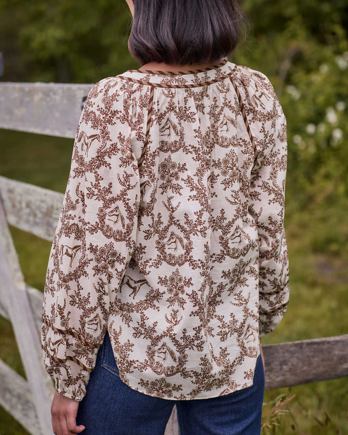 Back view of the oat Western Crown Kaia blouse showing the horse, floral, and horseshoe print, gathered yoke, and curved hem.