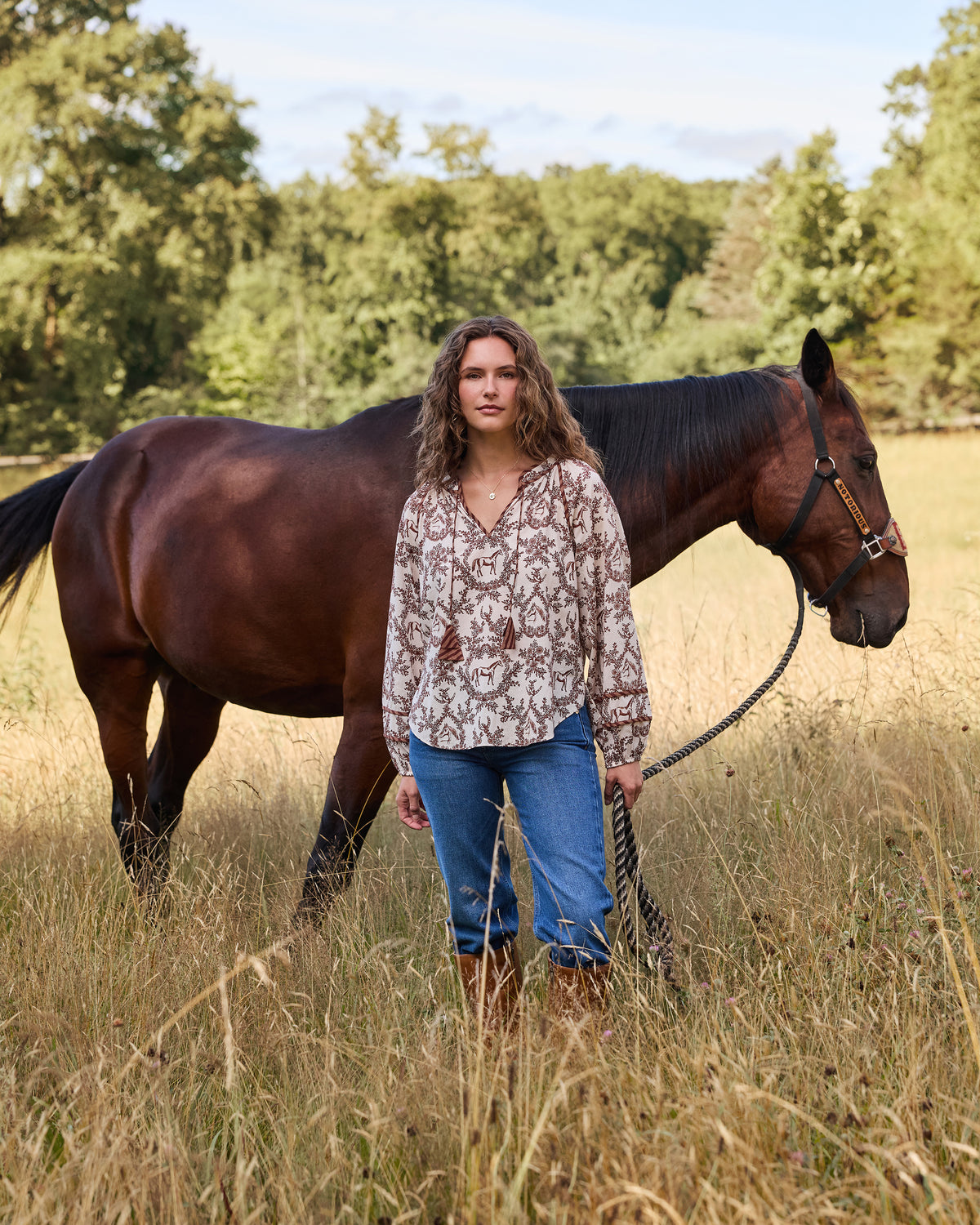 Person wearing the oat Western Crown Kaia blouse with a horse, floral, and horseshoe print beside a brown horse in a field.