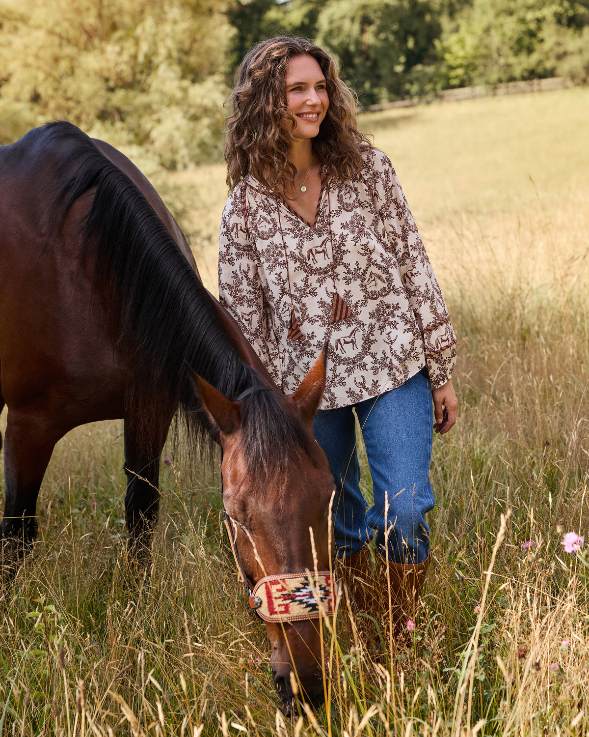 Person wearing the oat Western Crown Kaia blouse with horse, floral, and horseshoe print beside a horse in a grassy field.