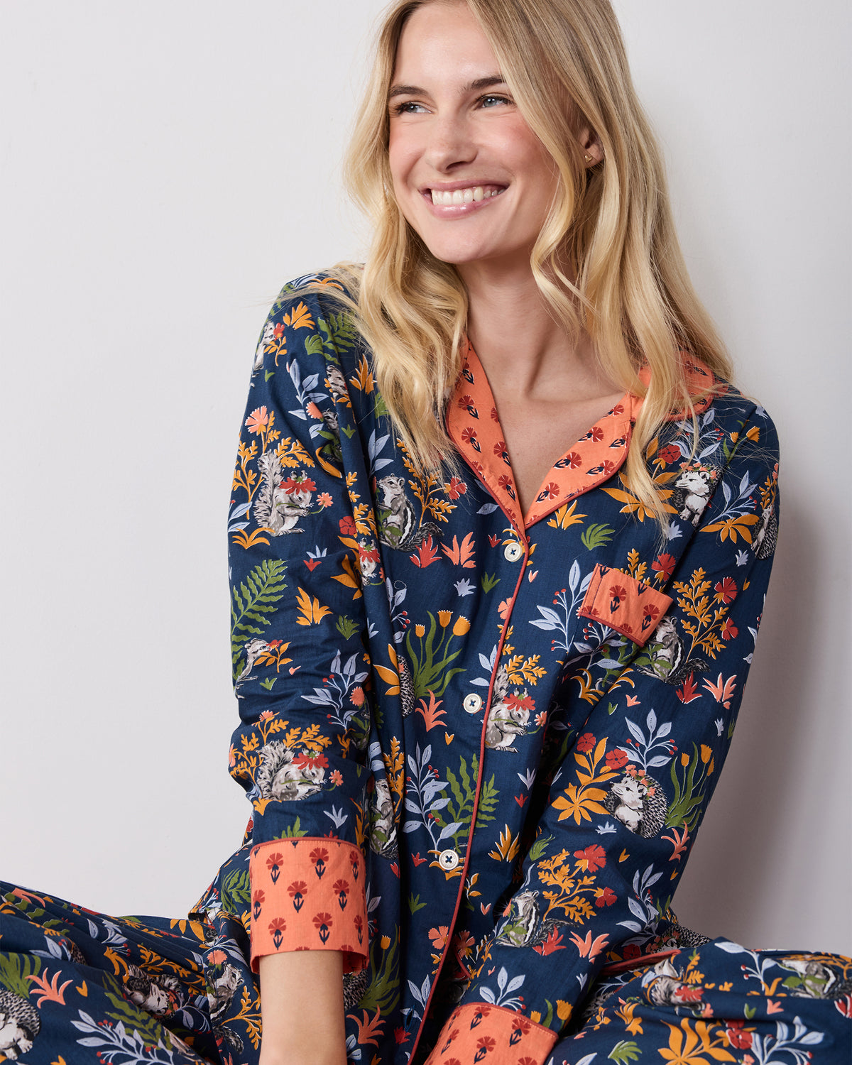 Seated close-up of the Woodland Critters indigo pajama top and matching pants, showing the coral collar, cuffs, buttons, and chest pocket.