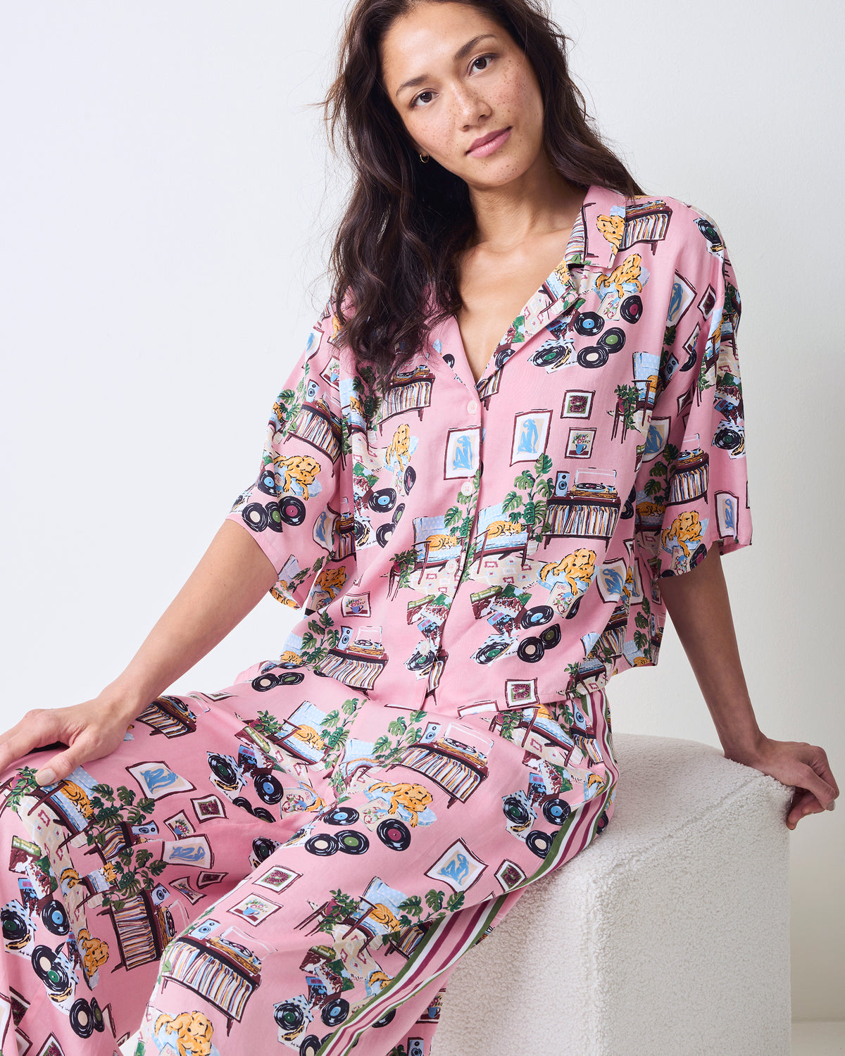 Front view of the petite pink Vinyl Collector pajama set worn while seated, showing the button-front cropped top, wide-leg pants, side stripe, and record-themed print.
