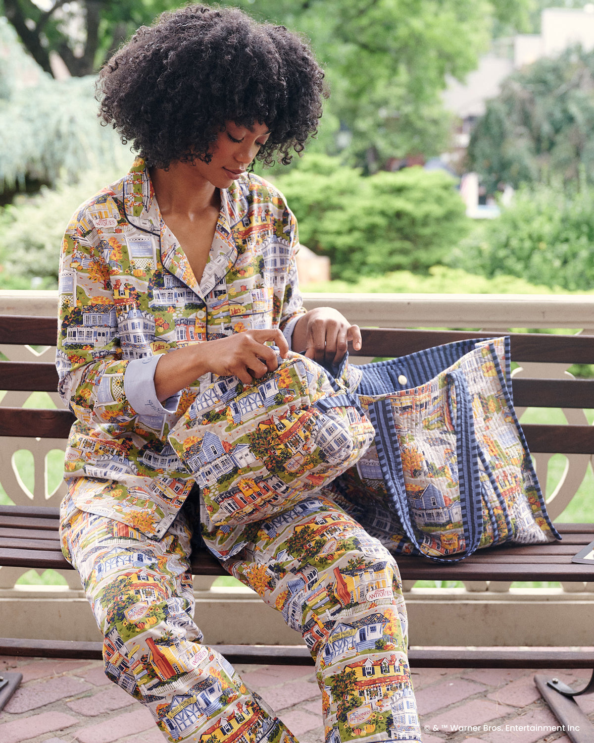 Model seated on a bench handling the Stars Hollow printed traveler tote, with the townscape pattern and blue checked lining visible.