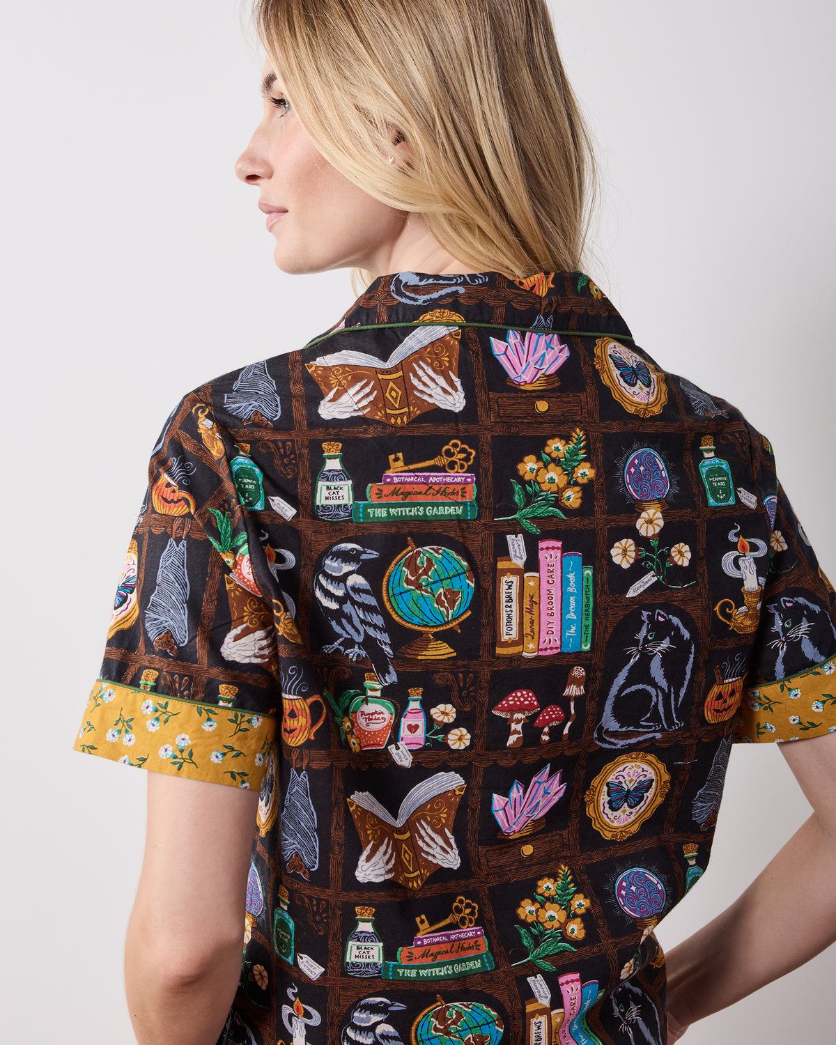 Back view of the Enchanted Study short pajama top, with short sleeves, a button front, and a dark cabinet-of-curiosities print featuring books, ravens, cats, bottles, candles, flowers, and other motifs.