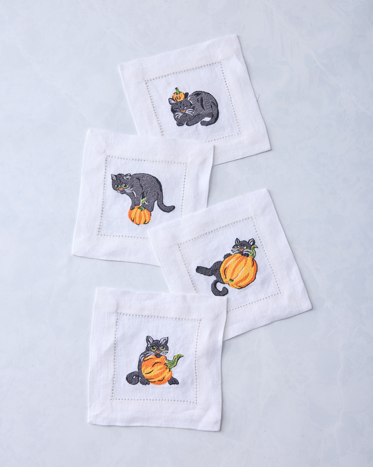 Four white cocktail napkins arranged together, embroidered with black cats and orange pumpkins.