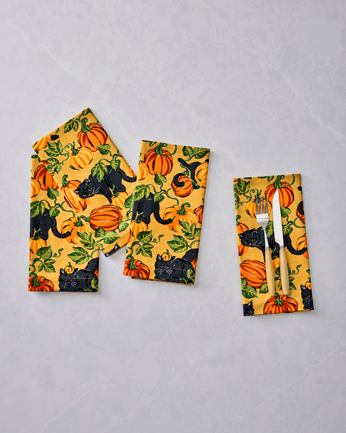 Three folded dinner napkins with an orange pumpkin-and-black-cat print; the right napkin holds a fork and knife.