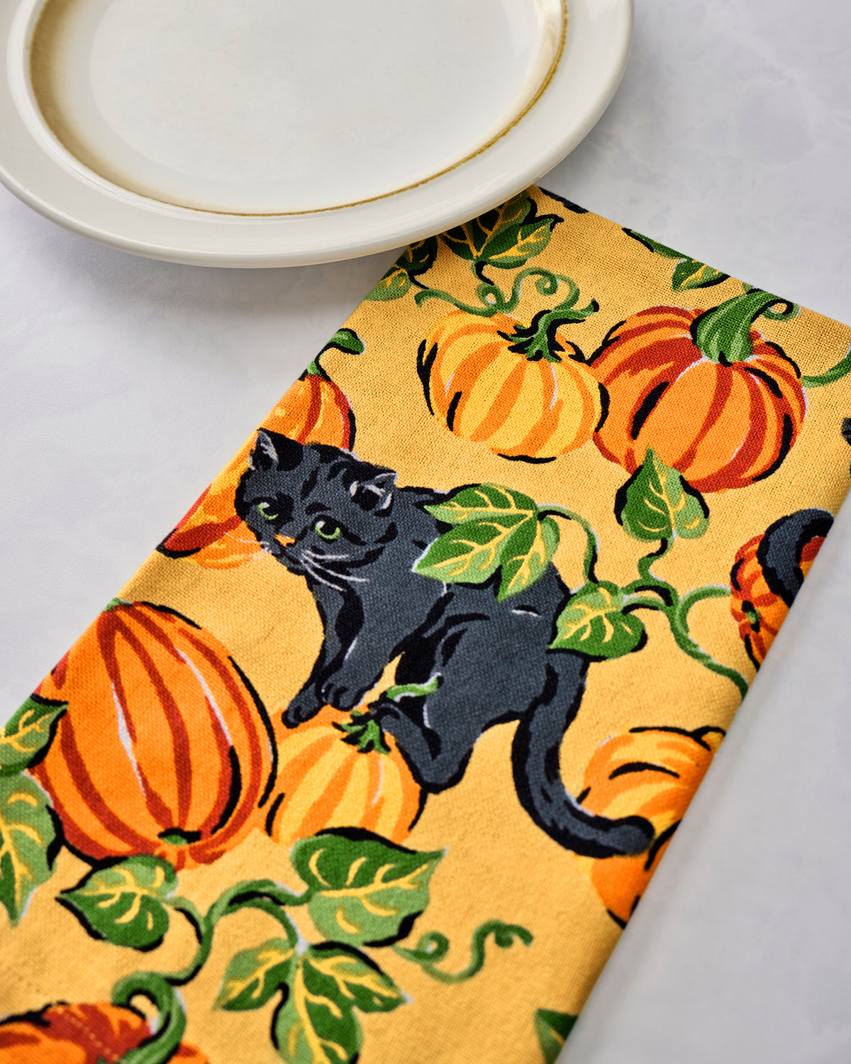 Close-up of a dinner napkin printed with orange pumpkins, green leaves, and a black cat beside stacked plates.