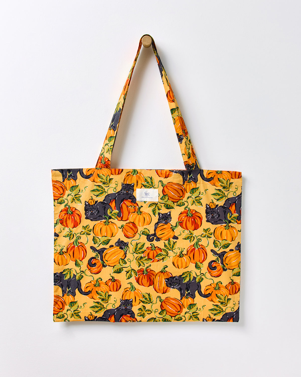 Pumpkin Prowl cotton tote hanging from a wall peg, covered in an orange pumpkin and black cat print with matching fabric handles.