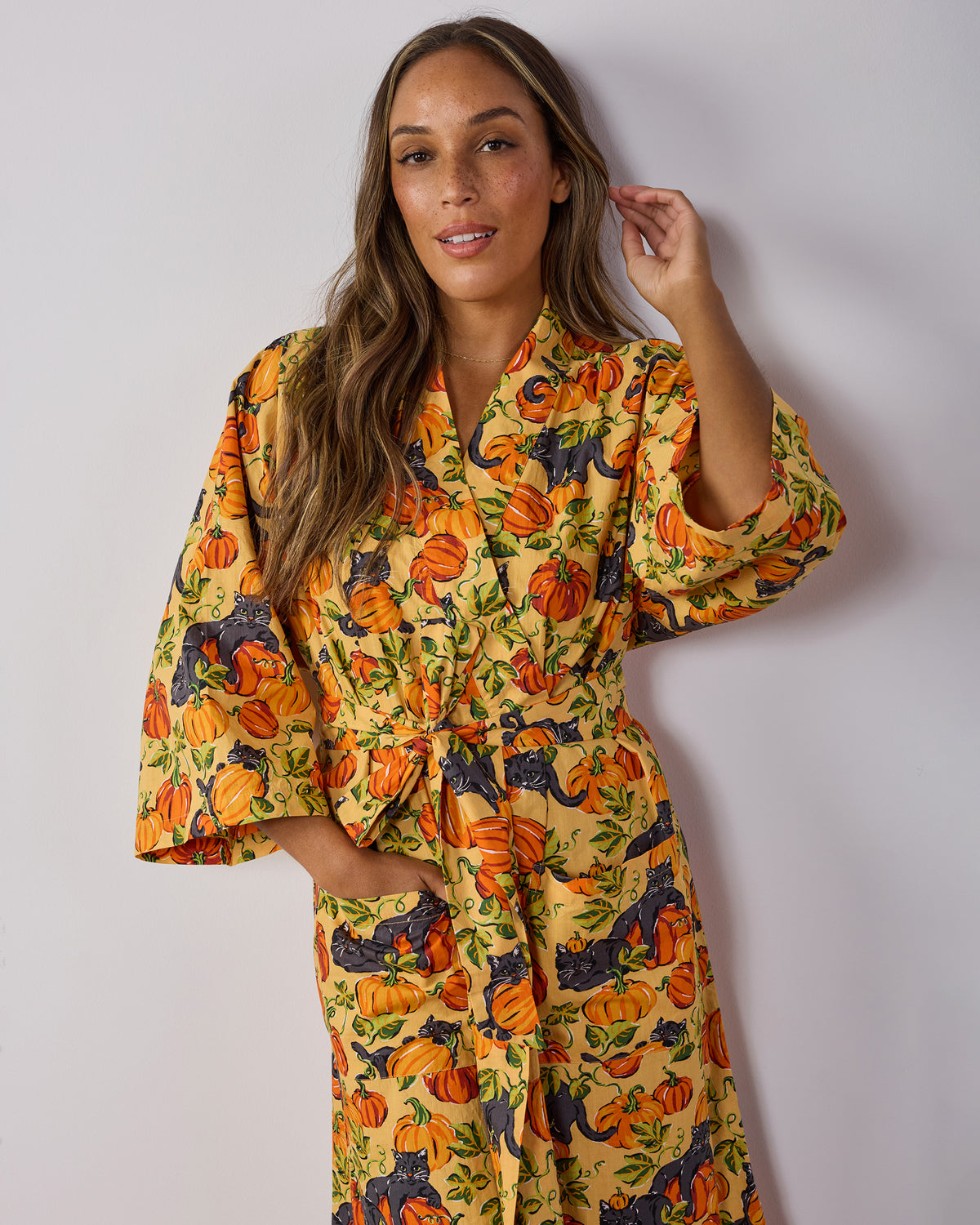 Front view of a person wearing the Pumpkin Prowl long robe with wide sleeves, tied belt, deep pockets, and a pumpkin and black-cat print.