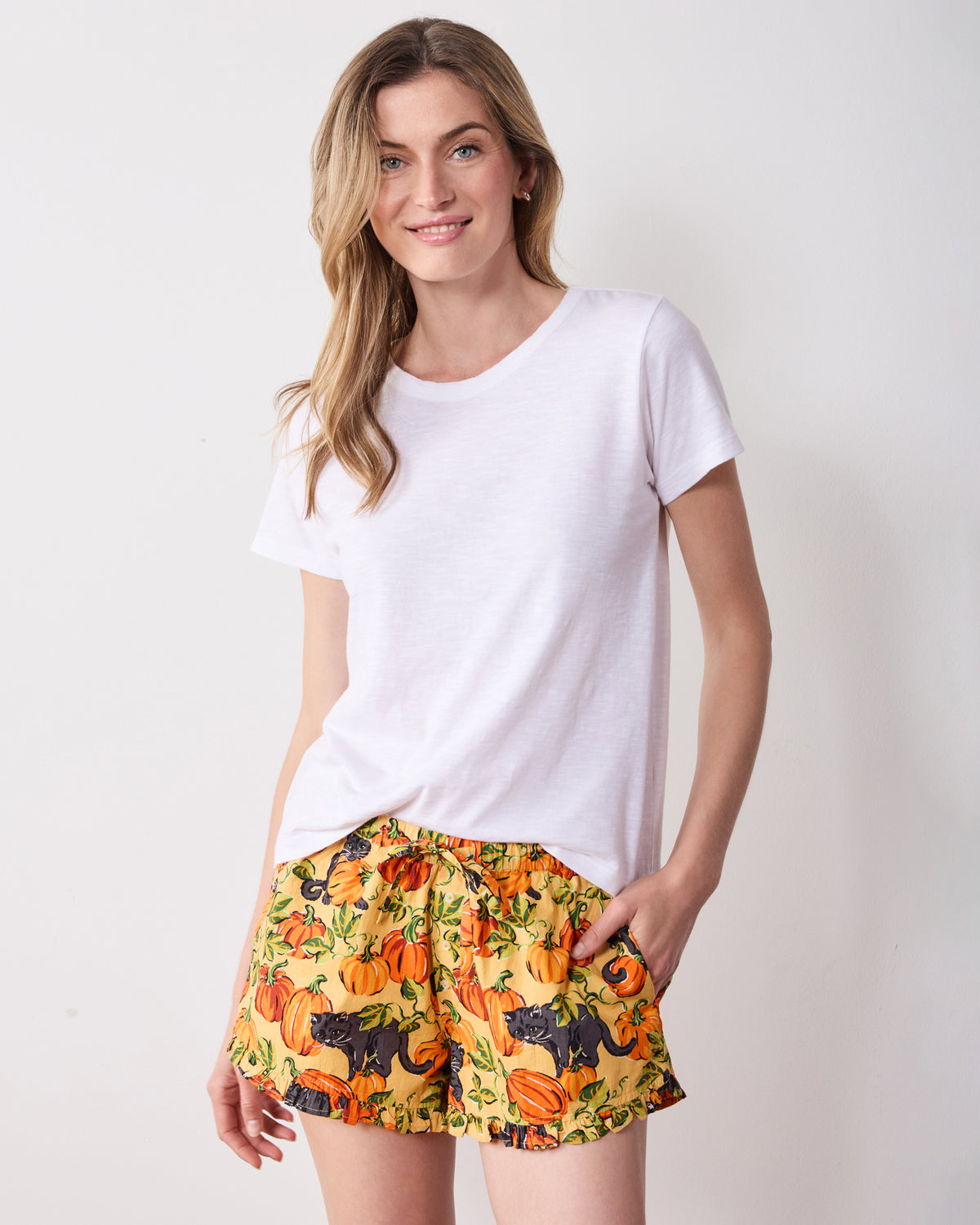 Front three-quarter view of Pumpkin Prowl salted caramel ruffle pajama shorts worn with a white T-shirt, showing the elastic waistband, pockets, and pumpkin-and-black-cat print.